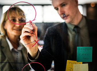 Two business colleagues collaborate on strategy, drawing a diagram with a red marker on a clear board.