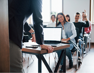 A speaker at a lectern with a laptop faces an engaged audience, with one person raising their hand to ask a question.