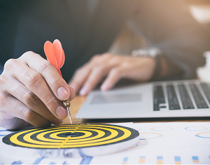 A professional places a red dart in the bullseye of a target on a desk, symbolizing achieving a business goal.