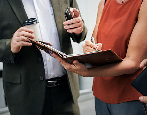 A woman takes notes in a planner held open by a male colleague who is holding a coffee cup and a smartphone.