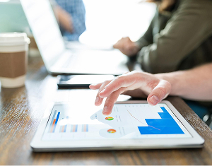 Close-up of a hand scrolling through business analytics charts and graphs on a tablet screen in a modern office.