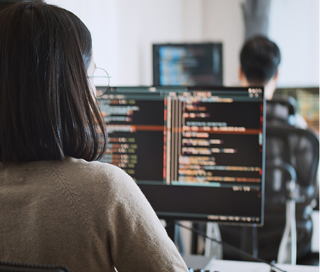 A female developer seen from behind, writing code on a computer screen with a colleague in the background.