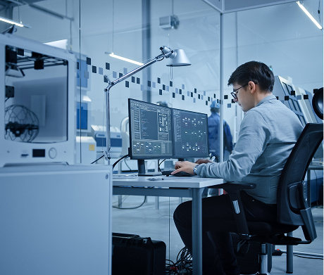An engineer in a modern laboratory works on a dual-monitor computer next to a 3D printer.