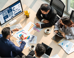 Overhead view of a business team in a meeting, analyzing data charts and a dashboard on a monitor.