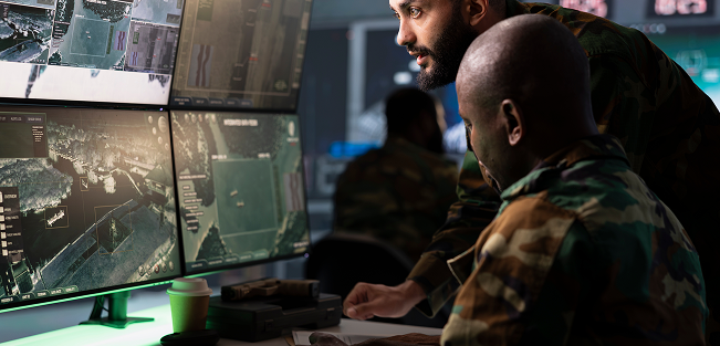 Two soldiers in camouflage monitor satellite images on multiple screens in a command center.